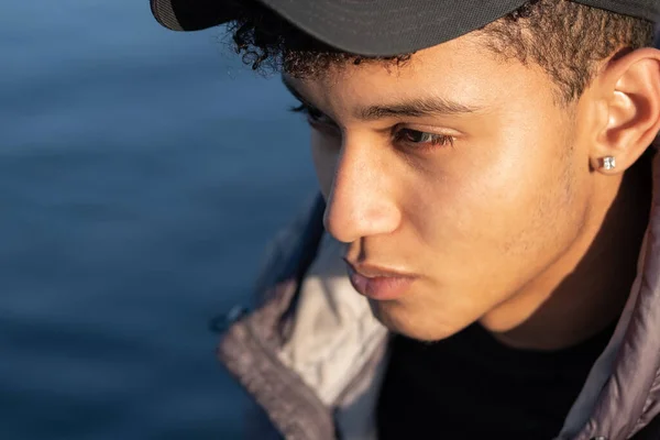Close-up view of a young man's face wearing a black cap. Man with tanned skin and curly hair. An earring in his ear. Details of the face. Thoughtful, deep thoughts, thinking.