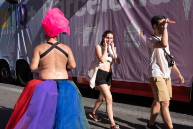 Barcelona, Spain - June 25, 2022: Celebration of Pride 2022 in Barcelona. Rear view of a reveler wearing a bizarre headdress and two tourists walking in the opposite direction along Avinguda del Paral-lel (Paral-lel Avenue). 