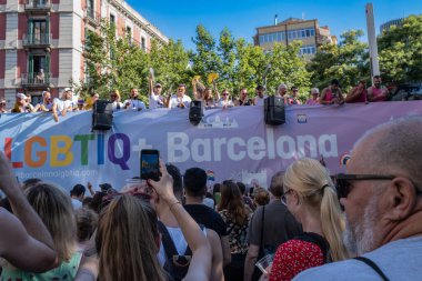 Barcelona, Spain - June 25, 2022: Pride 2022, crowd of people watching the parade. View from behind of a woman taking a photo with her smartphone as a float with the acronym LGBTIQ (Lesbian, Gay, Bisexual, Transgender, Intersex, Queer) passes by.