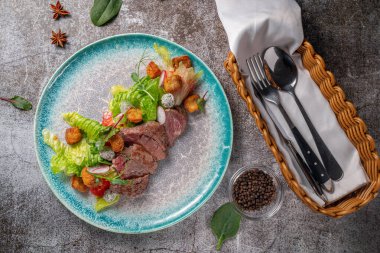 Serving a dish from the restaurant menu. Meat fried al dente (Semi-cooked) with vegetables, green salad and croutons on a plate against the background of a gray stone table, a delicious appetizer