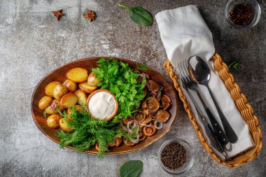 Serving a dish from a restaurant menu: country-style baked potatoes with pickled mushrooms and onions, cream sauce, dill and parsley greens on a plate against the background of a gray stone table