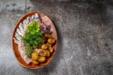 Serving a dish from a restaurant menu. Salted sliced herring fish with pickled mushrooms and onions, cream sauce, dill and parsley greens on a plate against the background of a gray stone table