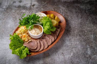 Serving a dish from the restaurant menu. Sliced beef tongue with pickles, cabbage, sauces and herbs on a plate against a gray stone table, a delicious appetizer