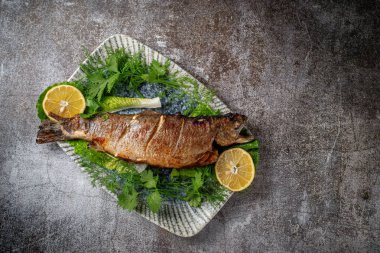 Roasted Whole Trout with lemon, lettuce and dill and parsley greens on a plate against a gray stone table 