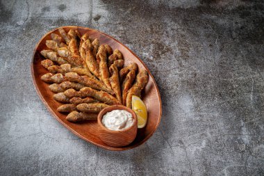 An appetizer in a restaurant, fried sprat on a wooden plate with lemon and cream sauce against a gray stone table 