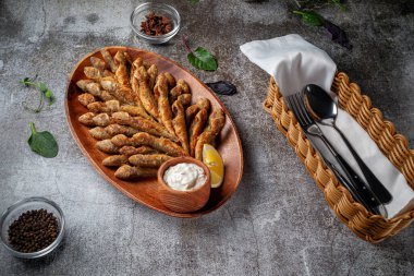 An appetizer in a restaurant, fried sprat on a wooden plate with lemon and cream sauce against a gray stone table 