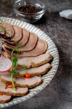 An appetizer in a restaurant, assorted meats. Sliced meat and sausage in layers on a plate with greens against a gray stone table 