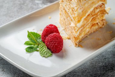 Piece of layered cream cake with raspberries and mint in a white plate against a gray stone table in a restaurant, a delicious breakfast 