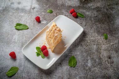 Piece of layered cream cake with raspberries and mint in a white plate against a gray stone table in a restaurant, a delicious breakfast 
