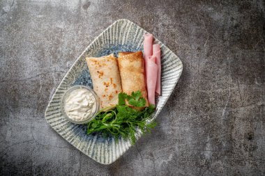 Pancakes filled with ham in a plate with parsley and dill greens and sour cream on a gray stone table, flatla