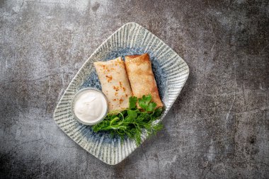 Stuffed pancakes in a plate with parsley and dill greens and sour cream on a gray stone table