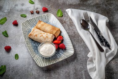 A delicious breakfast. Sweet fried pancakes filled with strawberries in a white plate with sour cream on a gray stone table with a napkin and cutlery