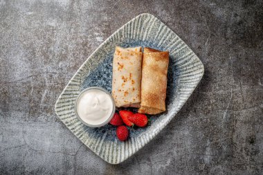 A delicious breakfast. Sweet fried pancakes filled with strawberries in a white plate with sour cream on a gray stone table with a napkin and cutlery