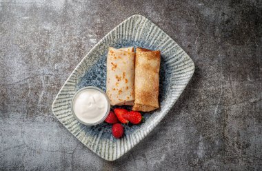 A delicious breakfast. Sweet fried pancakes filled with strawberries in a white plate with sour cream on a gray stone table with a napkin and cutlery