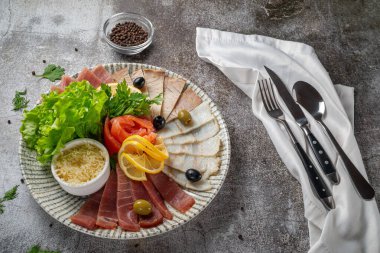 An appetizer in a restaurant, assorted meats. Sliced meat and sausage in layers on a plate with greens against a gray stone table 