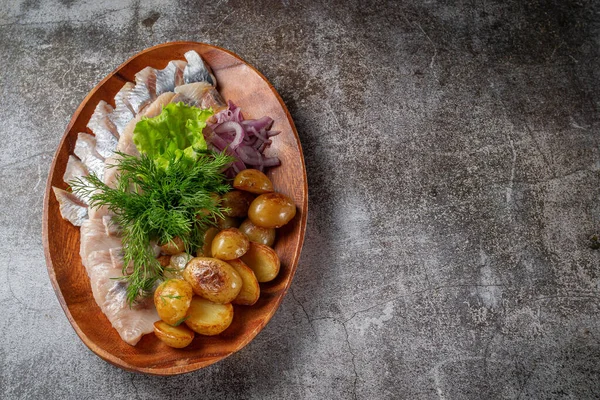 Serving a dish from a restaurant menu. Salted sliced herring fish with pickled mushrooms and onions, cream sauce, dill and parsley greens on a plate against the background of a gray stone table