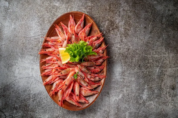 An appetizer in a restaurant, boiled shrimp on a wooden plate with lemon and herbs against a gray stone table