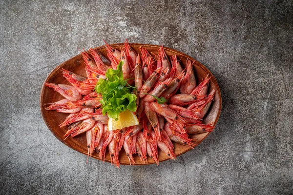 An appetizer in a restaurant, boiled shrimp on a wooden plate with lemon and herbs against a gray stone table