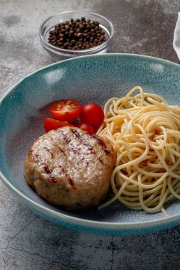 Spaghetti pasta with meat patty and fresh tomatoes on a blue plate against a gray stone table. 