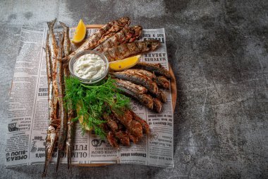 An appetizer in a restaurant, fried sprat on a wooden plate with lemon and cream sauce against a gray stone table 
