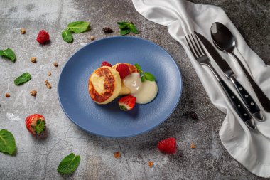 A delicious breakfast. Sweet fried pancakes with raspberries, strawberries and mint in a blue plate with sour cream on a gray stone table