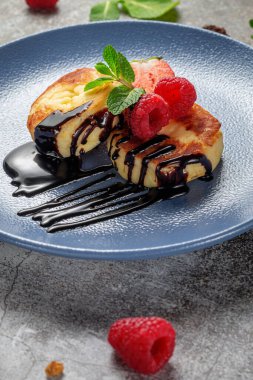 Fried pancakes with chocolate syrup and raspberries and mint in a blue plate against a gray stone table. A sweet breakfast in the morning 
