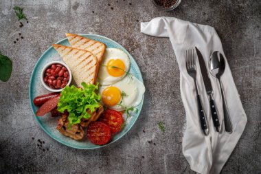 Serving a dish from the restaurant menu. Delicious and healthy breakfast with fried eggs, beans, sausages, grilled vegetables, tomatoes and mushrooms with salad on a plate against a gray stone table