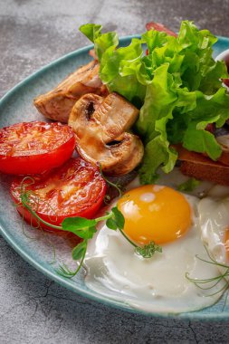 Serving a dish from the restaurant menu. Delicious and healthy breakfast with fried eggs, beans, sausages, grilled vegetables, tomatoes and mushrooms with salad on a plate against a gray stone table