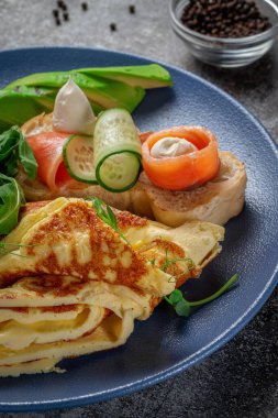 Omelet with salmon, fresh herbs and tomatoes in a blue plate with spices against a gray stone table