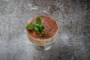Classic tiramisu dessert with mint leaves in a glass, with cocoa and chocolate in the background of a gray stone table 