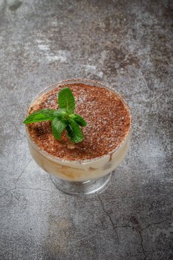 Classic tiramisu dessert with mint leaves in a glass, with cocoa and chocolate in the background of a gray stone table 