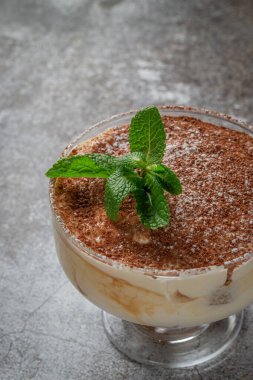 Classic tiramisu dessert with mint leaves in a glass, with cocoa and chocolate in the background of a gray stone table 