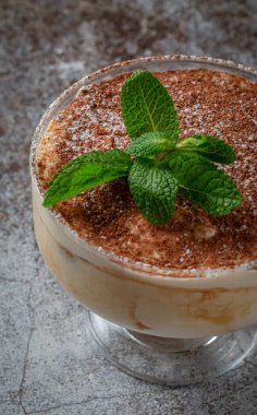 Classic tiramisu dessert with mint leaves in a glass, with cocoa and chocolate in the background of a gray stone table 