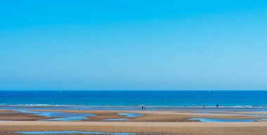 People on beach walking along shore of ocean with low tide and background of clear blue sky.