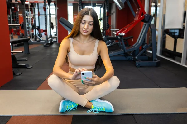 Akıllı telefonlu atletik kız spor salonunda mindere oturur.