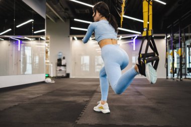 Woman doing squats weighted lunges exercise for butt legs with suspension straps. TRX exercise in gym
