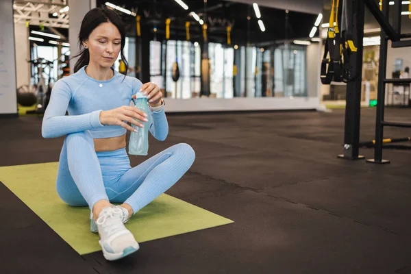 Spor salonundaki paspasın üzerine oturup bir şişe su açan sportif kadın.
