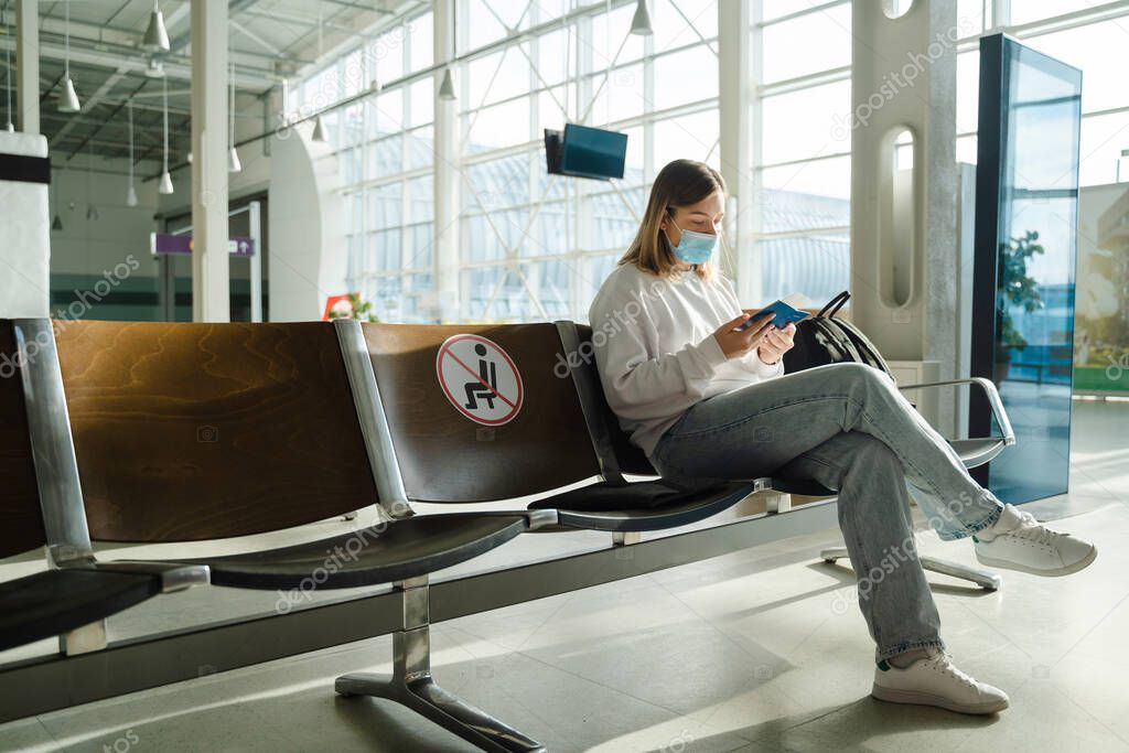 Female tourist in mask sitting in airport terminal lounge on empty marked chairs under new normal and covid social distance regulations. Woman sitting in airport lounge and looking at her passport and travel document