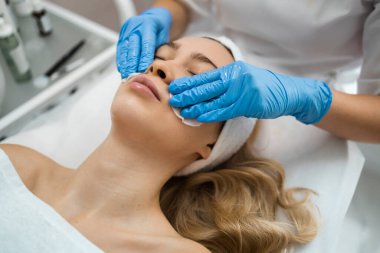 Hands of professional beautician wiping facial skin with cotton discs. Face of female client with closed eyes lying on couch and enjoying process during cosmetic procedure in cosmetology salon