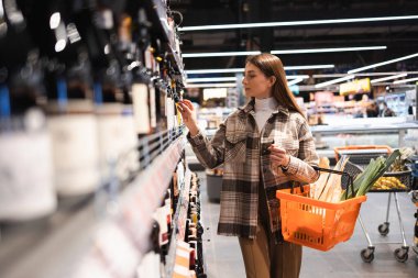 Young woman with shopping basket looking at racks with wine in store. Girl chooses wine in the supermarket
