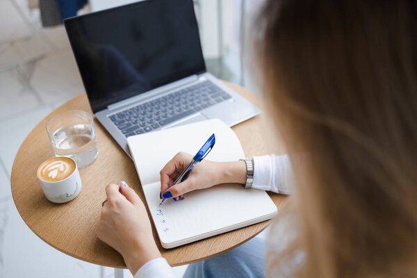 Неузнаваемая женщина сидит за столом в кафе и пишет в блокноте. Чашка кофе, стакан воды и ноутбук на столе у фрилансера