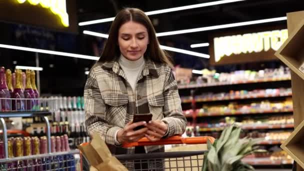 Bella Giovane Donna Utilizza Smartphone Mentre Cammina Con Carrello Supermercato — Video Stock