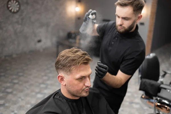 美容師は理容室で椅子に座って男性のクライアントの髪にヘアスプレーを適用します — ストック写真