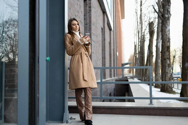 美しい若い女性がコーヒーとスマートフォンを持つ建物の近くに立っています — ストック写真