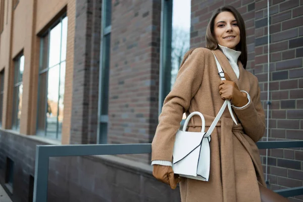 可愛いですCaucasian女の子でA茶色のコートとともに白い袋ポージング近くレンガ造りの建物 — ストック写真