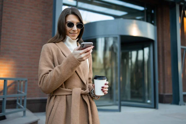 Stylische Frau Braunem Mantel Und Brille Mit Kaffeetasse Nutzt Smartphone — Stockfoto