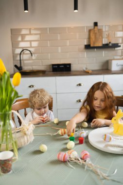 Kıvırcık saçlı oğlan ve kızıl saçlı kız kardeşi Paskalya yumurtalarını boyuyor.