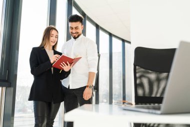 Genç sekreter patronuna bir defter gösteriyor. Ofiste iki şirket çalışanı.