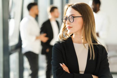 Kendine güveni tam bir iş kadını portresi. Kollarını kavuşturmuş ve iş arkadaşları arkasından iş çeviriyor.