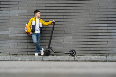 Elektrikli scooterlı çocuk tuğla duvarın yanında duruyor.